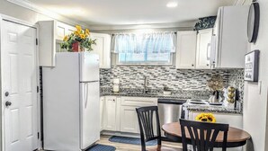 Una cocina compacta con armarios blancos, lavavajillas integrado, refrigerador, una mesa de comedor con sillas y una ventana con persianas.