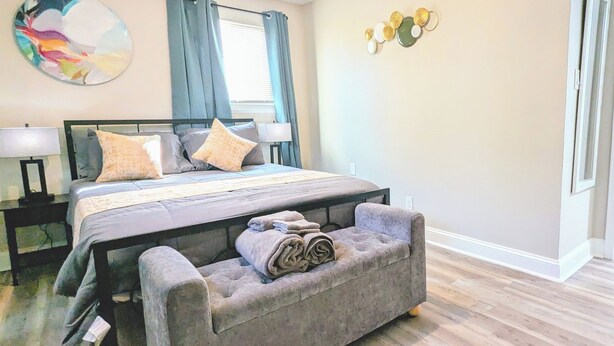 Un dormitorio con cama, mesitas de noche, un banco, una obra de arte en la pared y una ventana con cortinas.