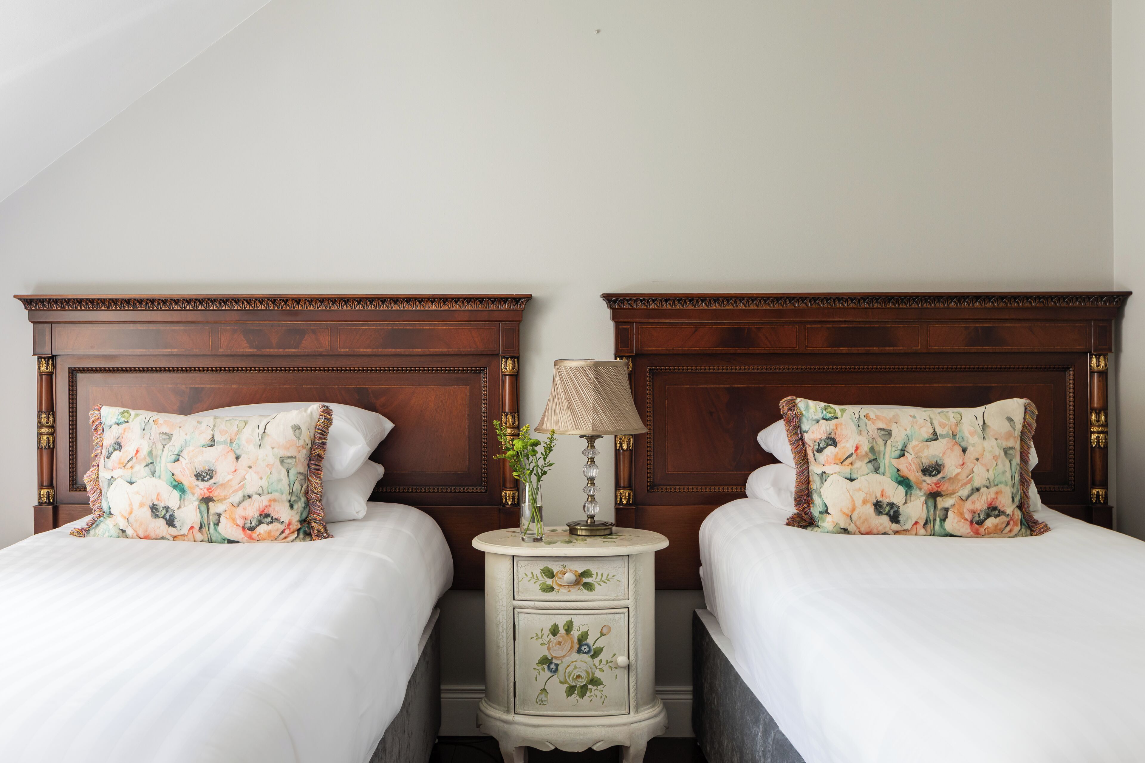 A double bed with floral-patterned pillows and a nightstand with a lamp and vase between the beds.