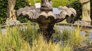 A stone fountain with water spouting from a statue, surrounded by greenery and a scenic lake view.