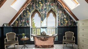 Una habitación con techo alto, una bañera grande, dos sillas, un aparador y un papel tapiz con motivos florales.
