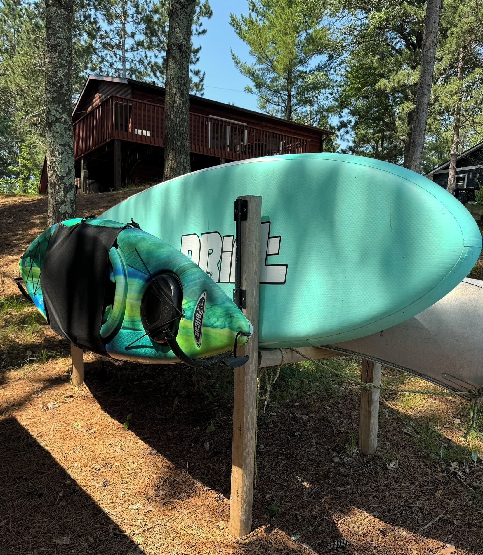 A kayak with a green and black design, secured to a wooden post in a wooded area with a cabin in the background.