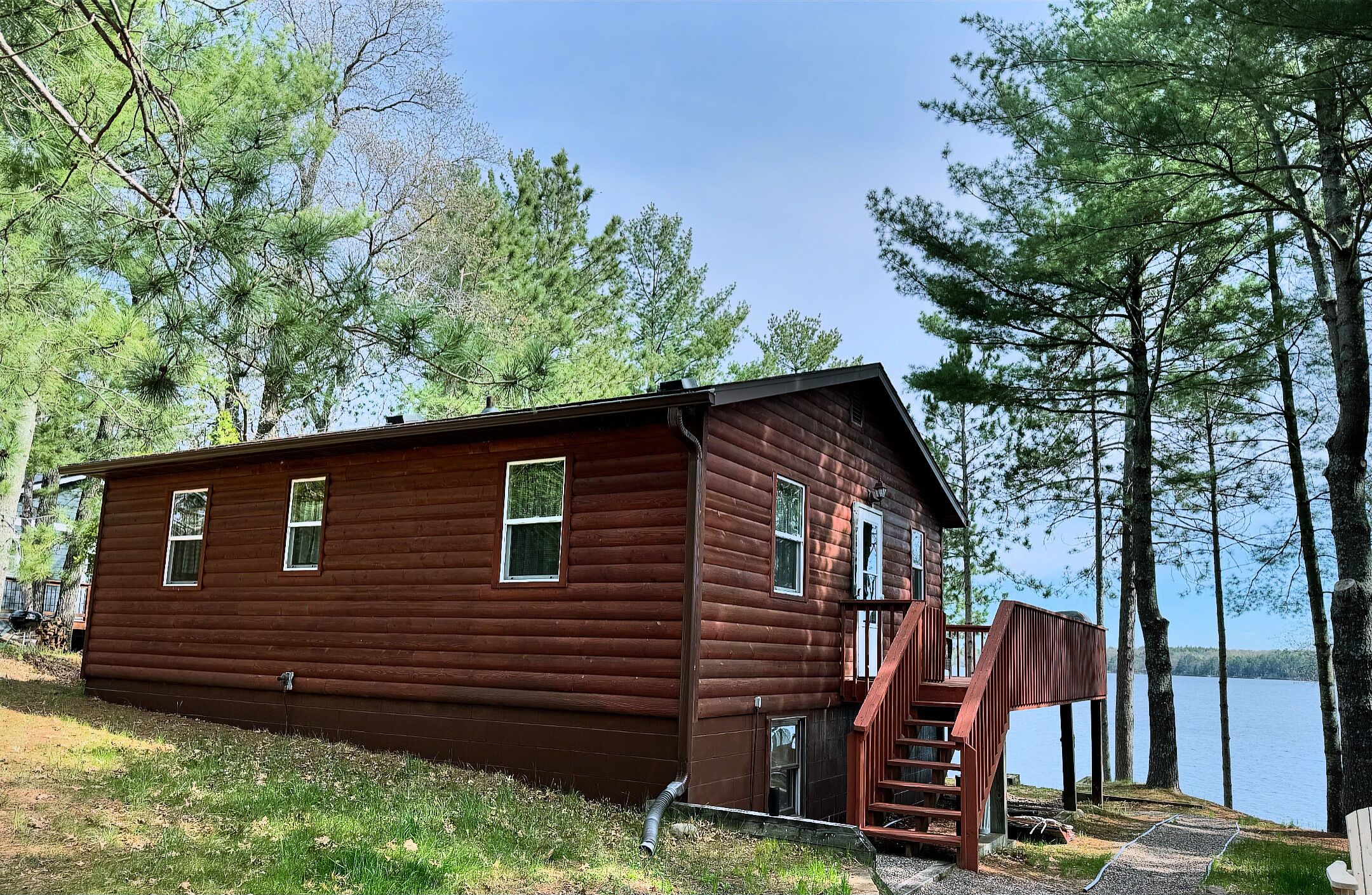 A wooden cabin with a staircase leading to a deck, surrounded by trees and a lake.