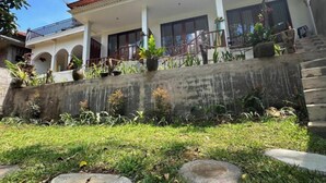 A two-story building with a tiled roof, a covered porch with potted plants, and a concrete retaining wall with a small waterfall feature. - Umah Yugadi Bali (Bali)