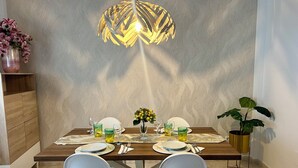 Un'area pranzo con tavolo in legno apparecchiato per due, sedie bianche, una lampada a sospensione decorativa e una parete con motivo.