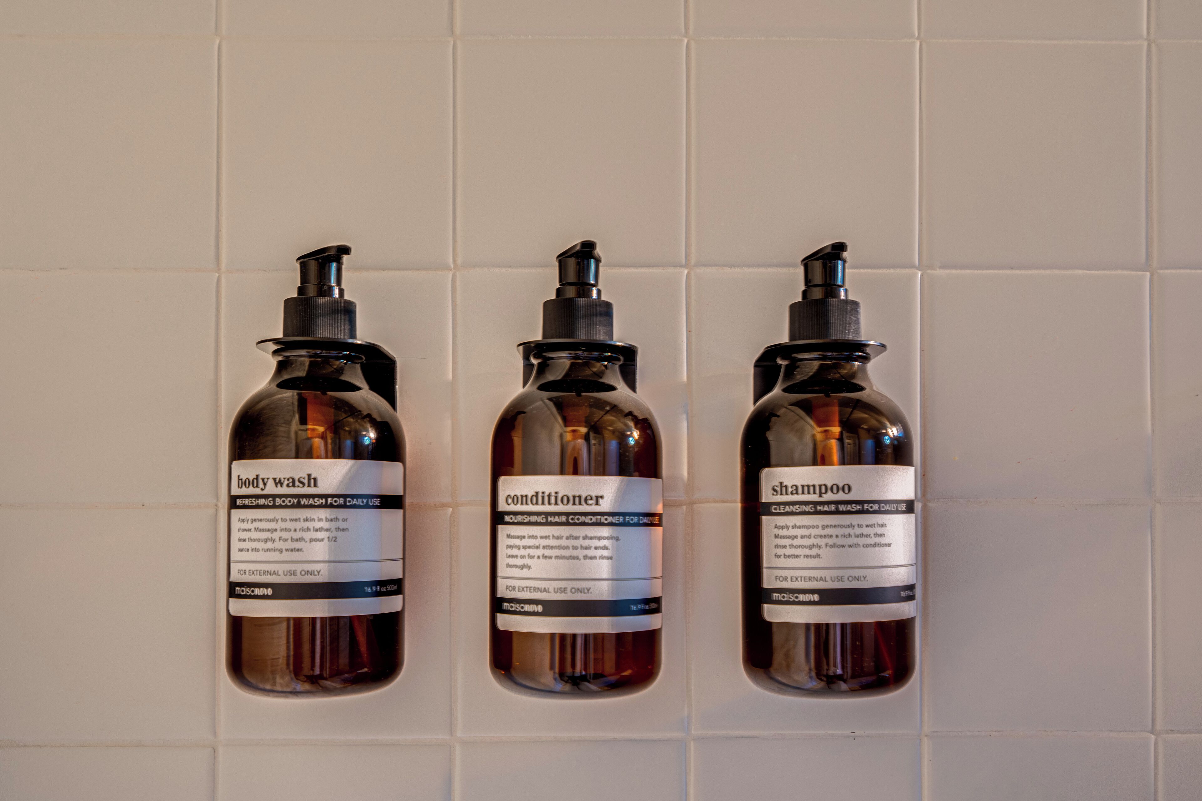 Tres botellas de jabón de cuerpo, acondicionador y champú dispuestas sobre una pared de baldosas.