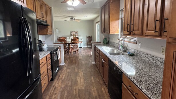 A kitchen with wooden cabinets, granite countertops, a black refrigerator, a ceiling fan, and a dining area with a table and chairs.
