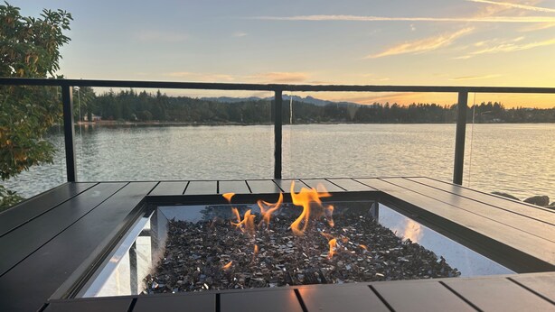 A rectangular outdoor fire feature with a metal frame, surrounded by a wooden deck, overlooking a body of water and distant mountains.