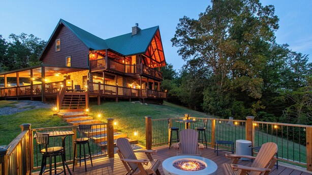 Una terraza de madera con mobiliario exterior, una chimenea y una casa grande con techo verde y balcones de madera.