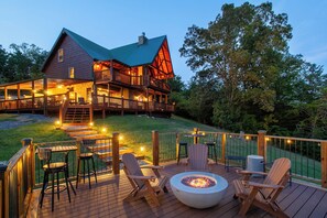 Eine Holzterrasse mit Außenmöbeln, einer Feuerstelle und einem großen Haus mit Gründach und hölzernen Balkonen.