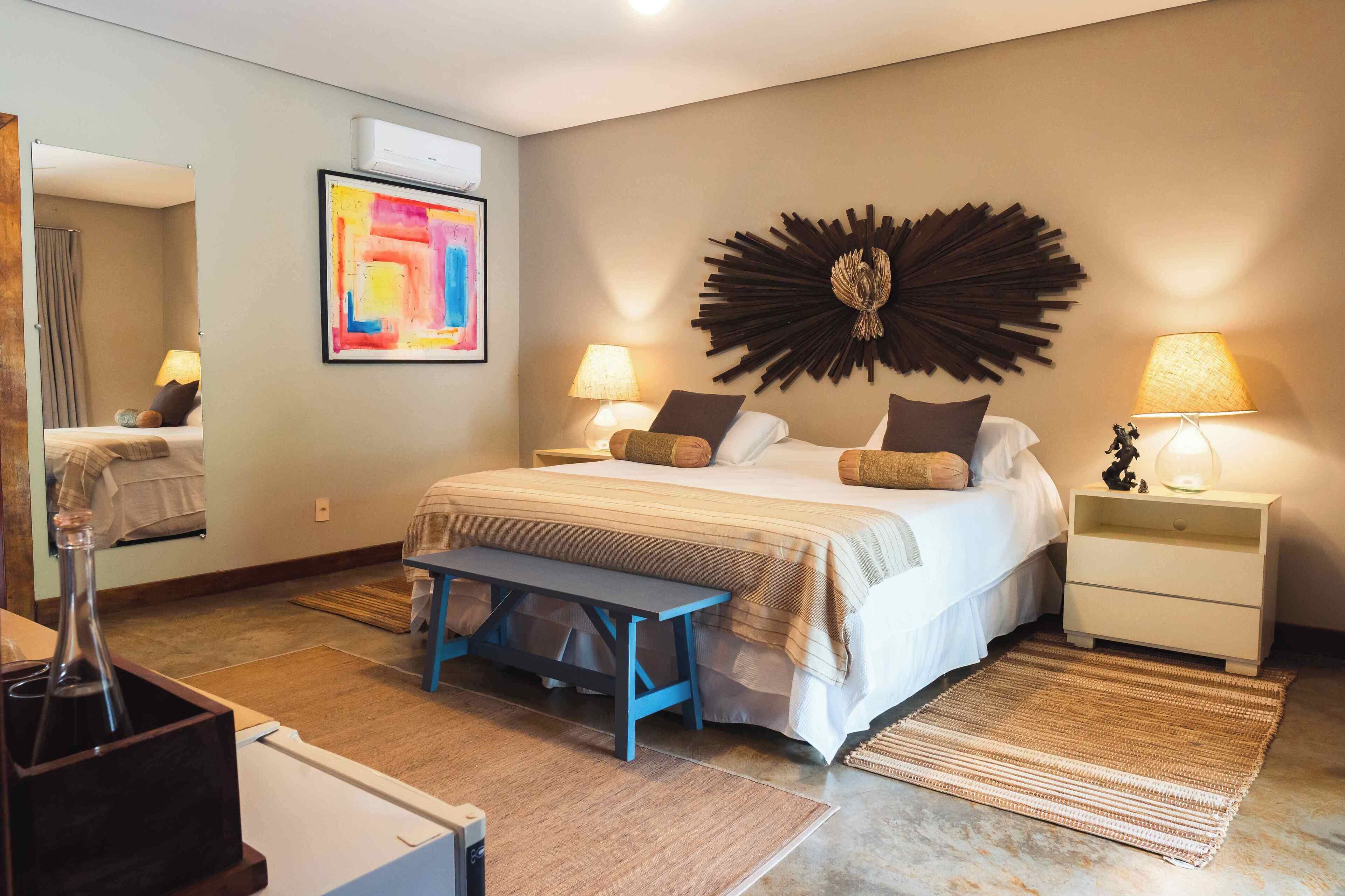 Una habitación de hotel moderna con una cama grande, mesitas de noche con lámparas, un banco, un espejo y un cuadro abstracto de colores.