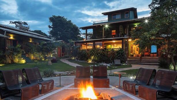 Un'area patio con un braciere circondato da poltrone relax, un giardino curato e un edificio a due piani illuminato con luci esterne.