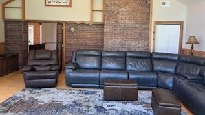 Een ruime woonkamer met een grote leren hoekbank, een kleinere leren fauteuil en een bijpassend poef. Er is een rustieke bakstenen accentmuur en een plafondventilator te zien.