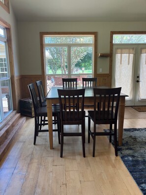 A dining area with a wooden table and chairs, large windows with a view of greenery, and a door with a curtain.