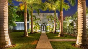 A pathway leading to a building with a waterfall feature, surrounded by palm trees and greenery.