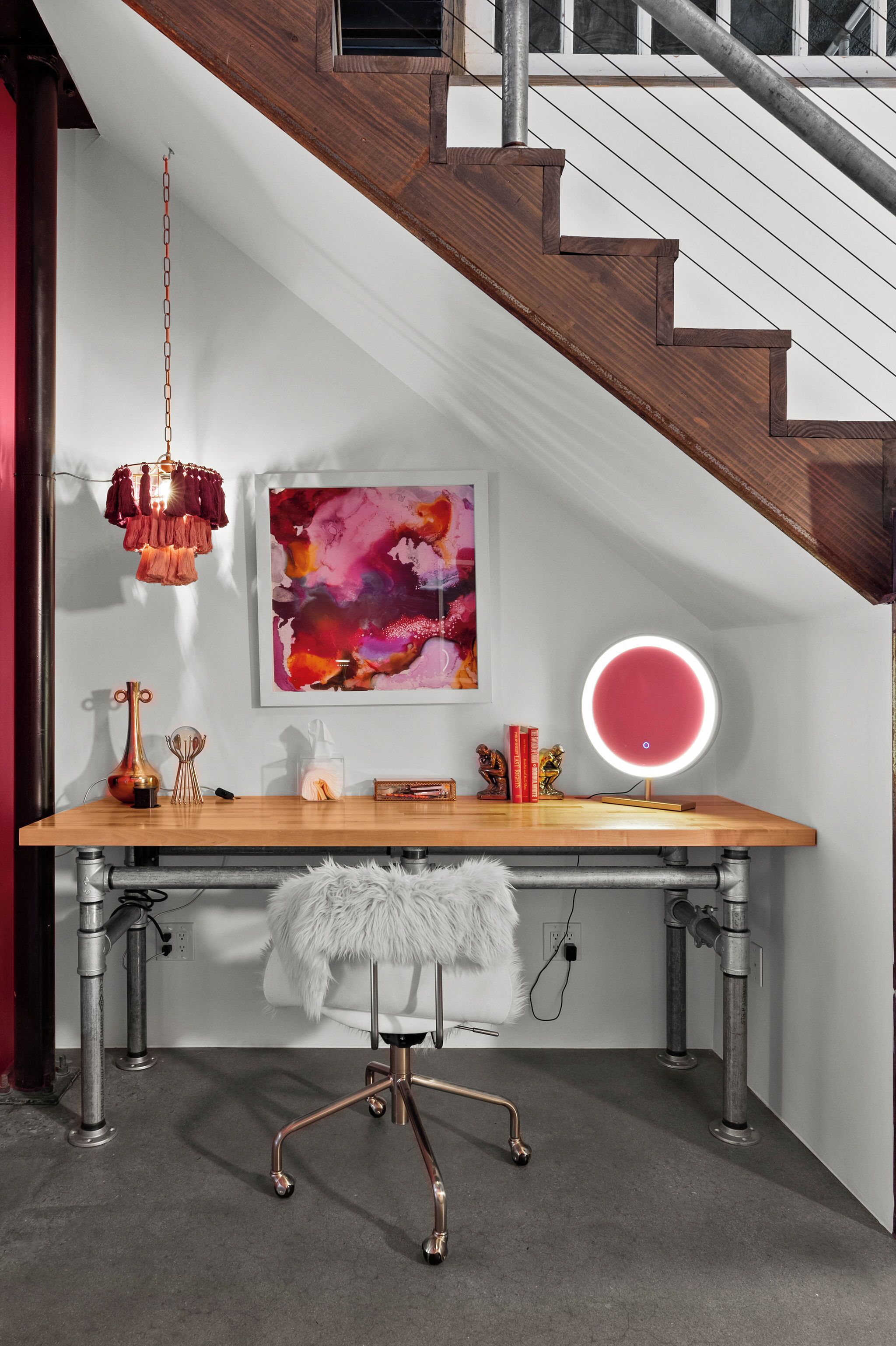 A modern workspace with a wooden desk, a white furry chair, a hanging light fixture, and a wall-mounted round mirror.