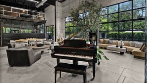 A spacious living room with a grand piano, comfortable seating, large windows, and a view of greenery.