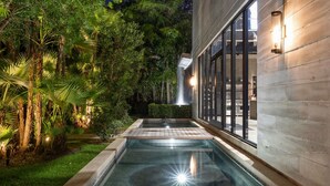 A swimming pool with a waterfall feature, surrounded by lush greenery and a modern building with large windows.