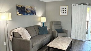 Ein Wohnzimmer mit grauem Sofa, einem kleinen Tisch und einem Sessel. Zwei Lampen und ein Bild hängen an der Wand.