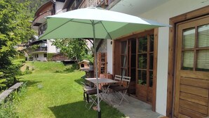 Un patio con una mesa y sillas bajo un paraguas grande, una puerta de madera y un jardín césped con un pequeño comedero para pájaros.