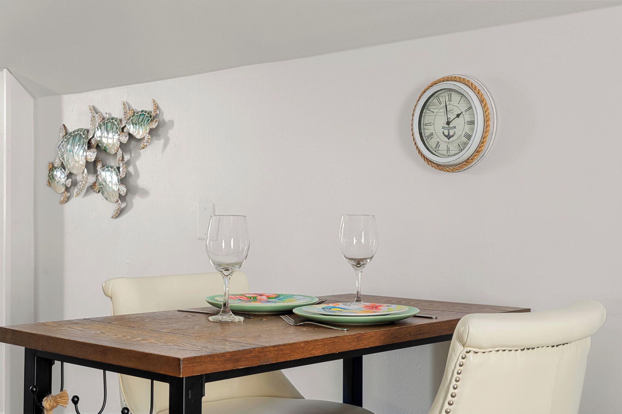 A dining table with two plates and two wine glasses, a wall clock, and a decorative wall piece.