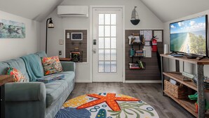 Ein gemütliches Wohnzimmer mit einem hellblauen Sofa, bunten Kissen, einem Teppich mit Seestern-Muster, einer Holz-TV-Bank und einem großen Fenster mit Blick aufs Meer.