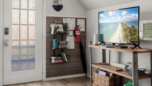 Ein Zimmer mit einem großen Fenster, einer Holzhilfe mit Büchern und einem Feuerlöscher, sowie einem Fernseher, der eine Strandansicht zeigt.