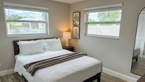 A neatly made bed with white bedding and a brown throw blanket, a bedside lamp, and a framed plant on the wall.