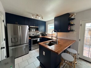 A modern kitchen with dark blue cabinets, stainless steel appliances, a wooden island with stools, and a view through a sliding glass door.