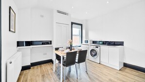 A modern kitchen with white cabinetry, a dining table with chairs, a built-in washing machine, and a large window. - MODERN DUPLEX APARTMENT LOCATED NEAR CITY CENTRE - FREE PARKING, WI FI & NETFLIX (Reading)