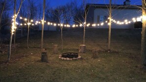 A rustic outdoor setting with a fire pit surrounded by trees, string lights, and a metal building in the background. - Copper Lodge - Lake Miltona
License #:  1999 (Miltona)