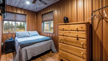 Un dormitorio con paredes de madera, una cama con colcha gris, un tocador de madera y dos ventanas con persianas.