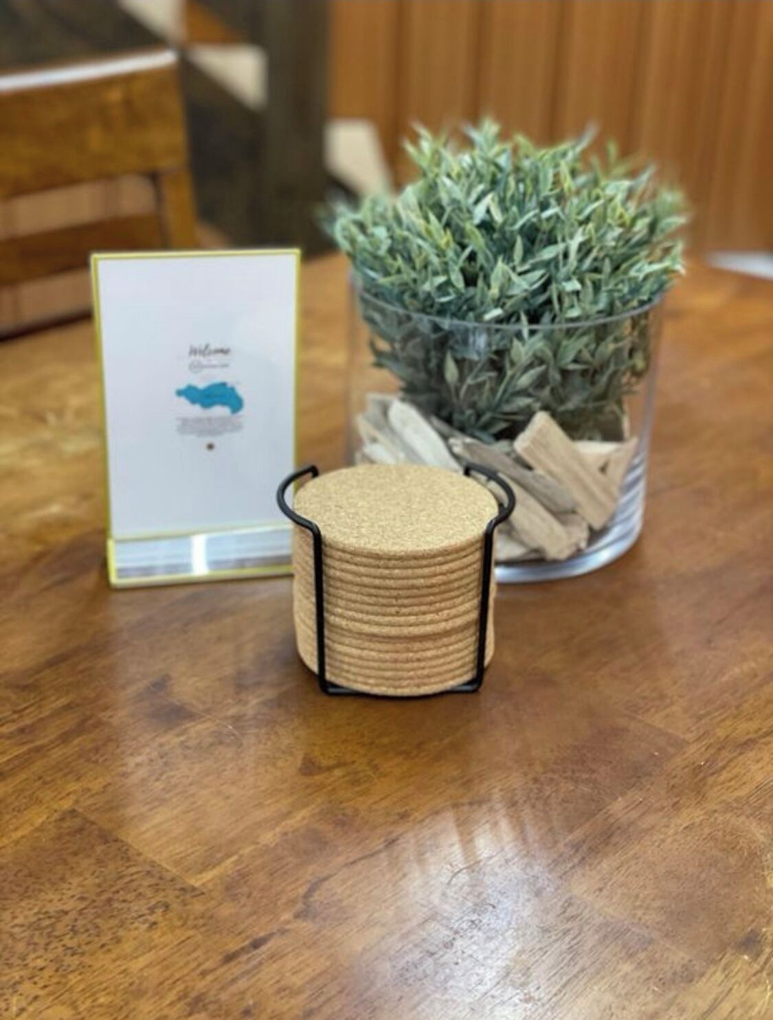 A wooden table with a framed welcome sign, a small round container with a black rim, and a glass vase with a green plant and wood pieces.