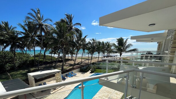 A modern hotel balcony with a pool, overlooking a beach with palm trees and the ocean.