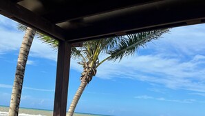 A wooden deck with a person lying on a hammock, overlooking a beach with palm trees and the ocean.