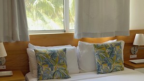 A bedroom with a bed, curtains, a nightstand, a lamp, and a window with a view of palm trees.