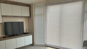 A living room with a sofa, a coffee table, a TV on a cabinet, and large windows with blinds.