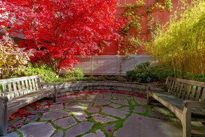 A courtyard with stone flooring, two wooden benches, and a variety of plants including a red-leafed tree.