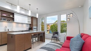 A modern kitchen with a central island, bar stools, and a red sofa with a grey pillow. - Sleek Kitchen Island and Garden Views (Victoria North)