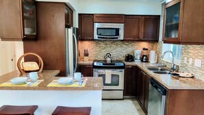 A modern kitchen with dark wood cabinets, stainless steel appliances, a granite countertop, and two wooden stools. - ☀️🌴Spinnaker 1312 by IRE Vacations - 2 bedroom Condo with Resort Amenities👙 (Oyster Pond)