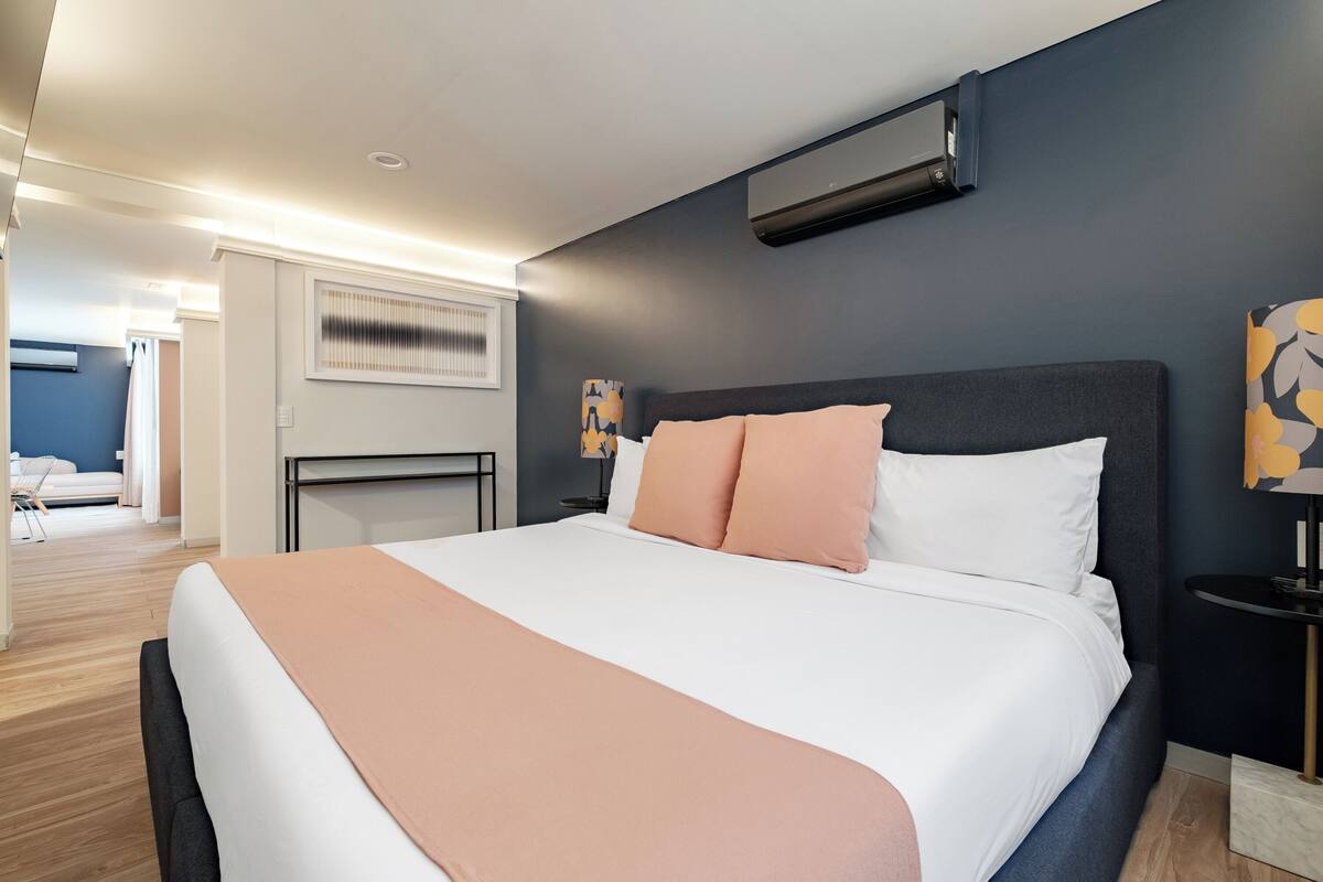 Een moderne hotelkamer met een groot bed, een nachtkastje, een wandairconditioner en uitzicht op de aangrenzende kamer.