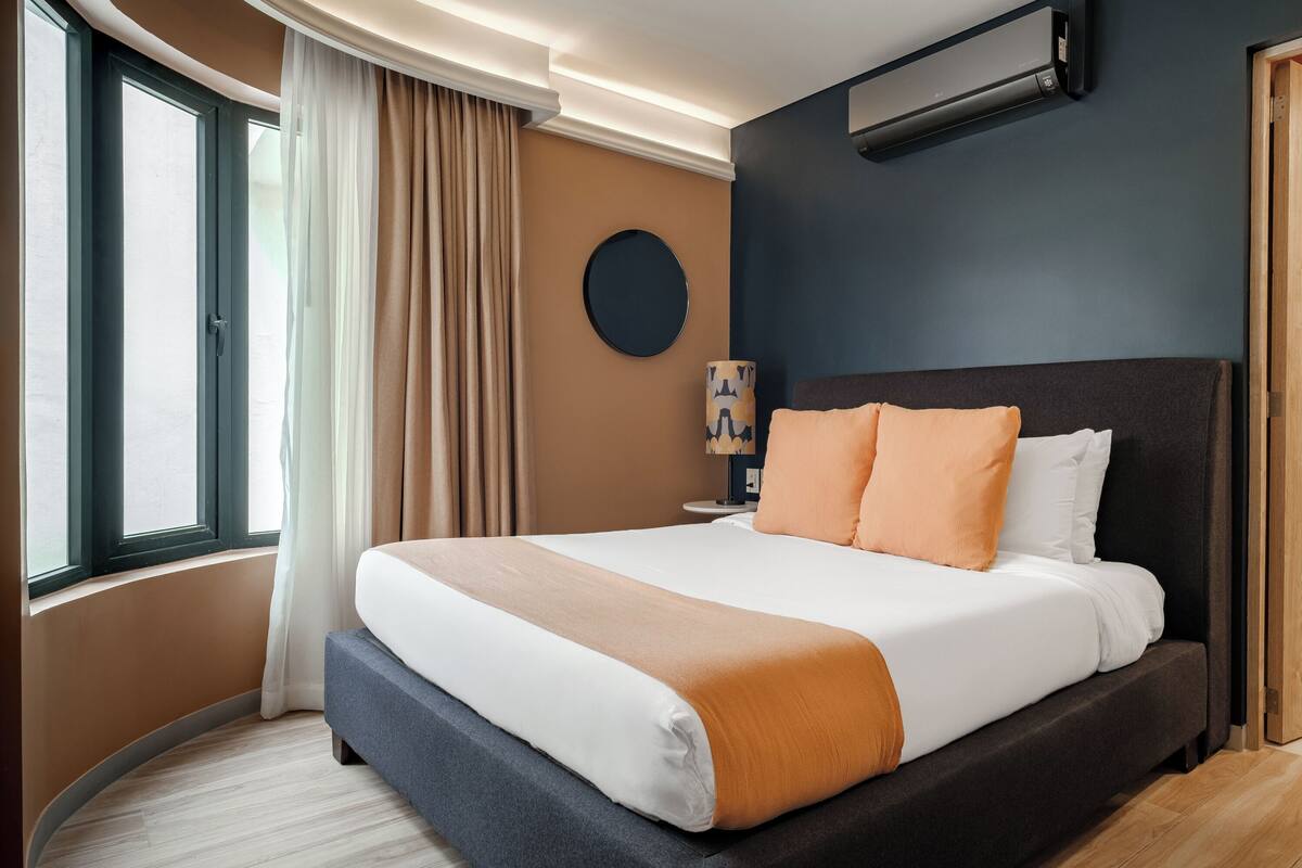 Een hotelkamer met een groot bed, een ronde wandklok, een nachtkastje met een lamp en een raam met gordijnen.