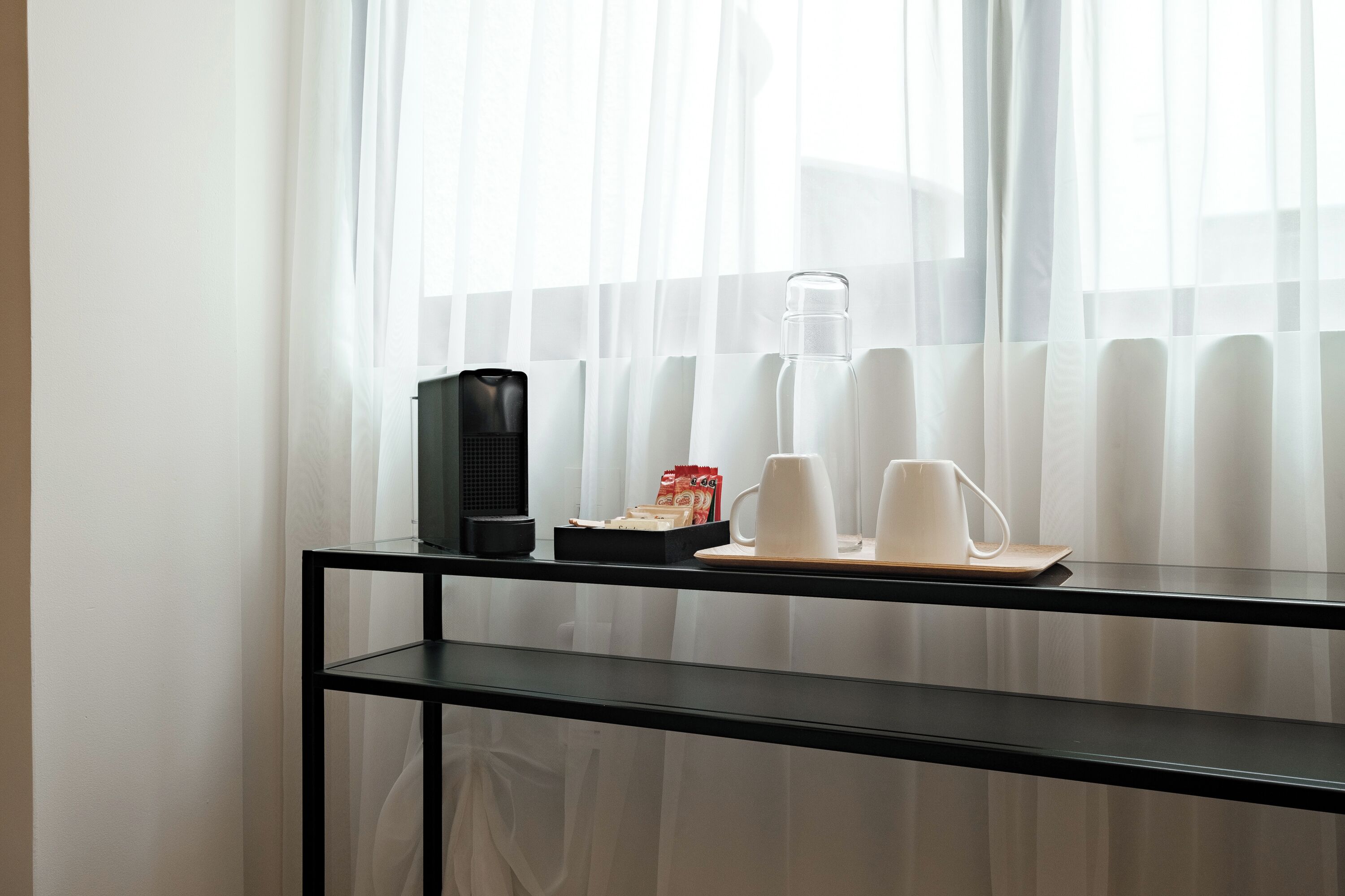 Una mesa de vidrio con una cafetera, tazas y una botella de agua cerca de una ventana con cortinas translúcidas.