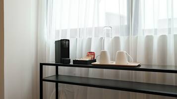 Una mesa de vidrio con una cafetera, tazas y una botella de agua cerca de una ventana con cortinas translúcidas.