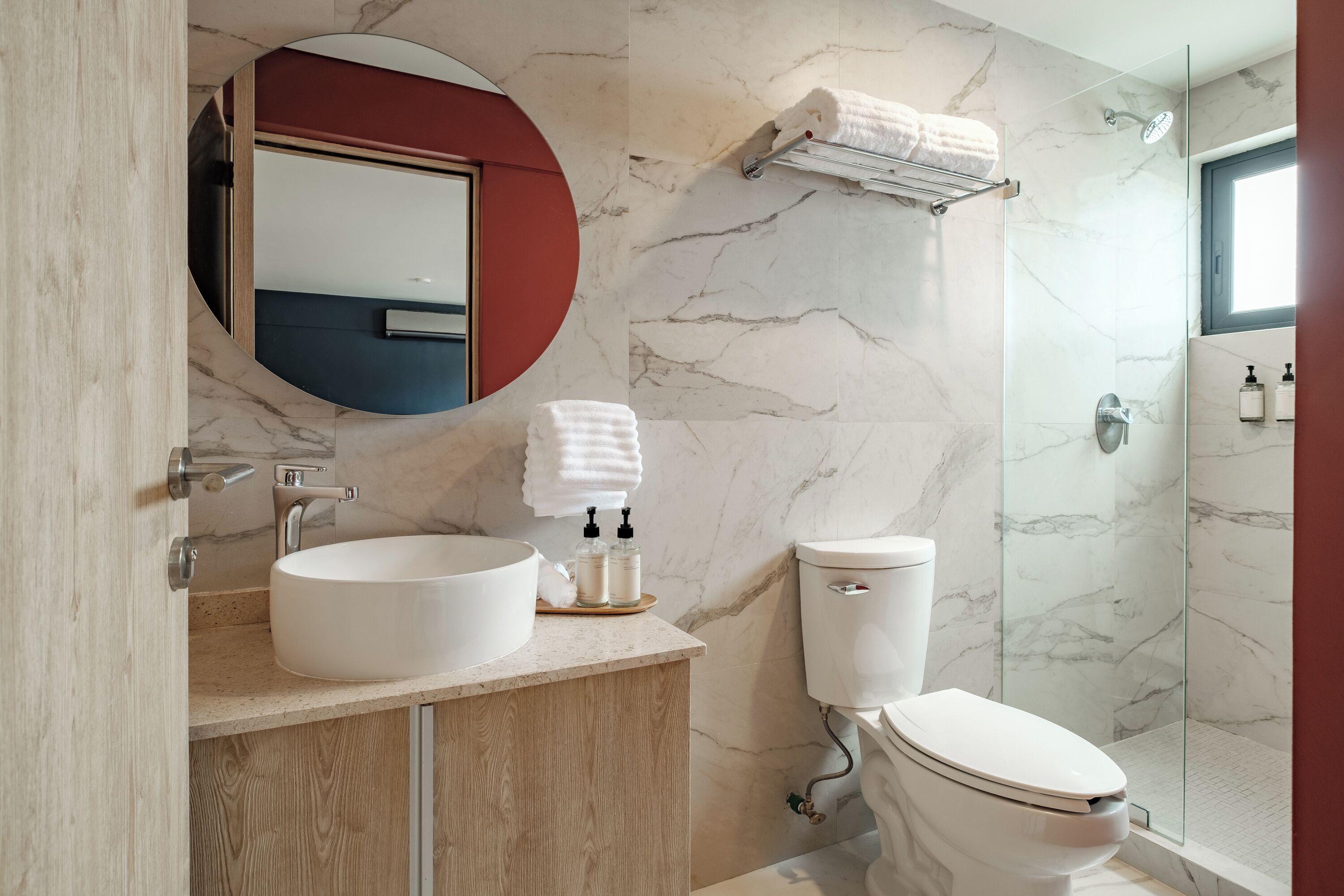 Ein modernes Badezimmer mit Marmorwand, einem runden Spiegel, einem Waschbecken mit Holzschrank, einer Toilette und einer Duschkabine aus Glas.