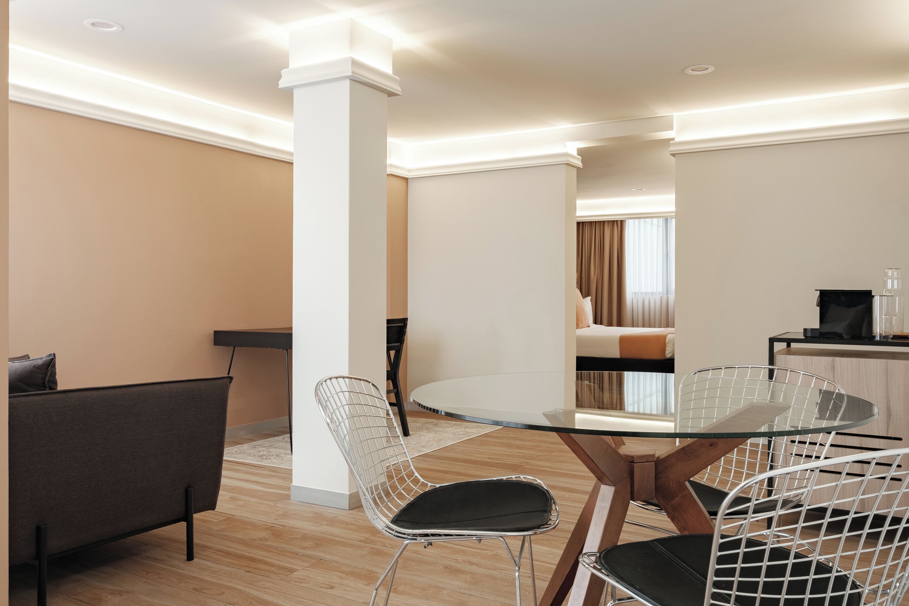 A modern dining area with a glass table and wire chairs, a sofa, and a kitchen area in the background.