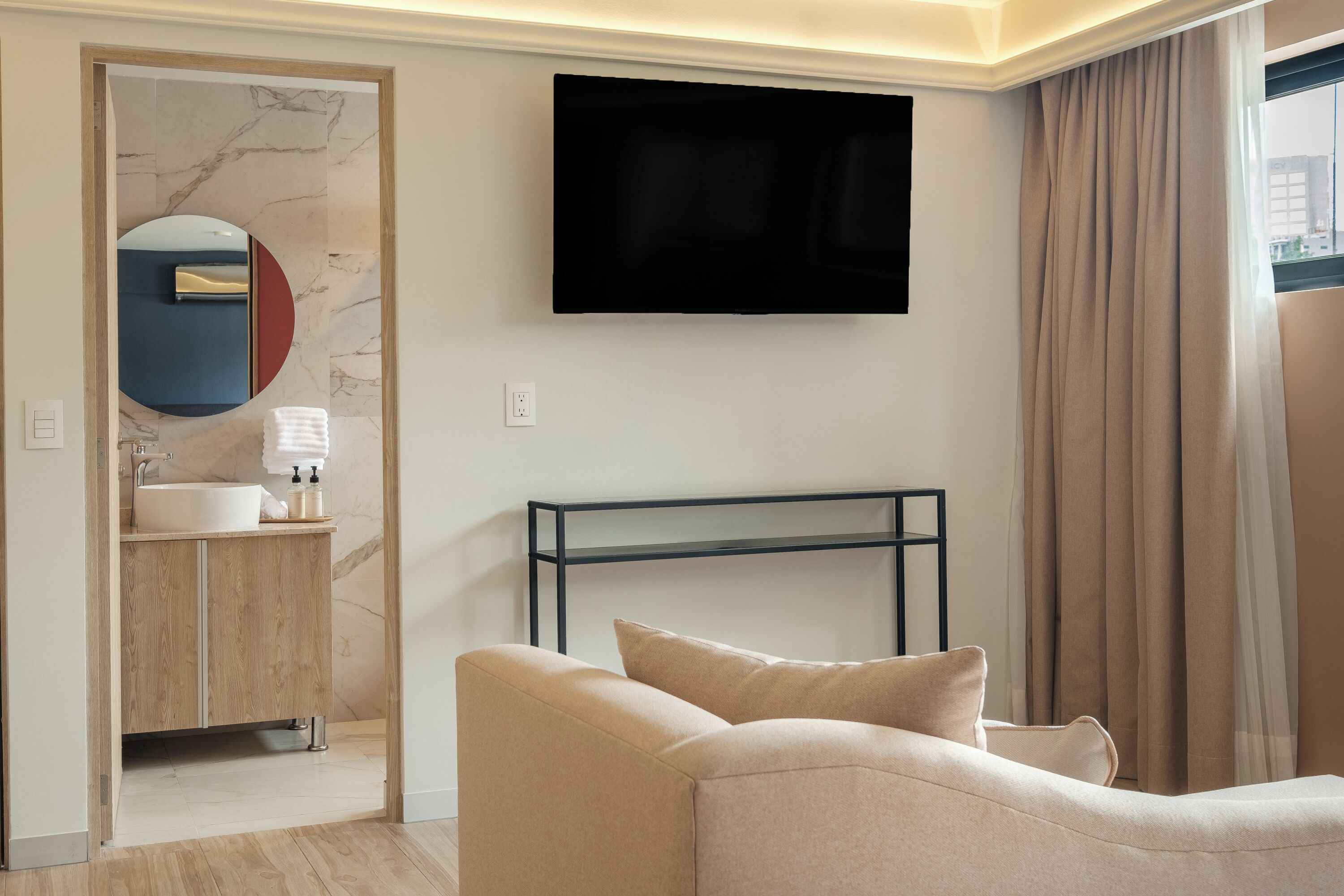 Habitació d'hotel moderna amb televisió de pantalla plana penjada a la paret, un tocador de fusta amb respatller de marbre, un sofà beix i un gran finestral amb cortines.