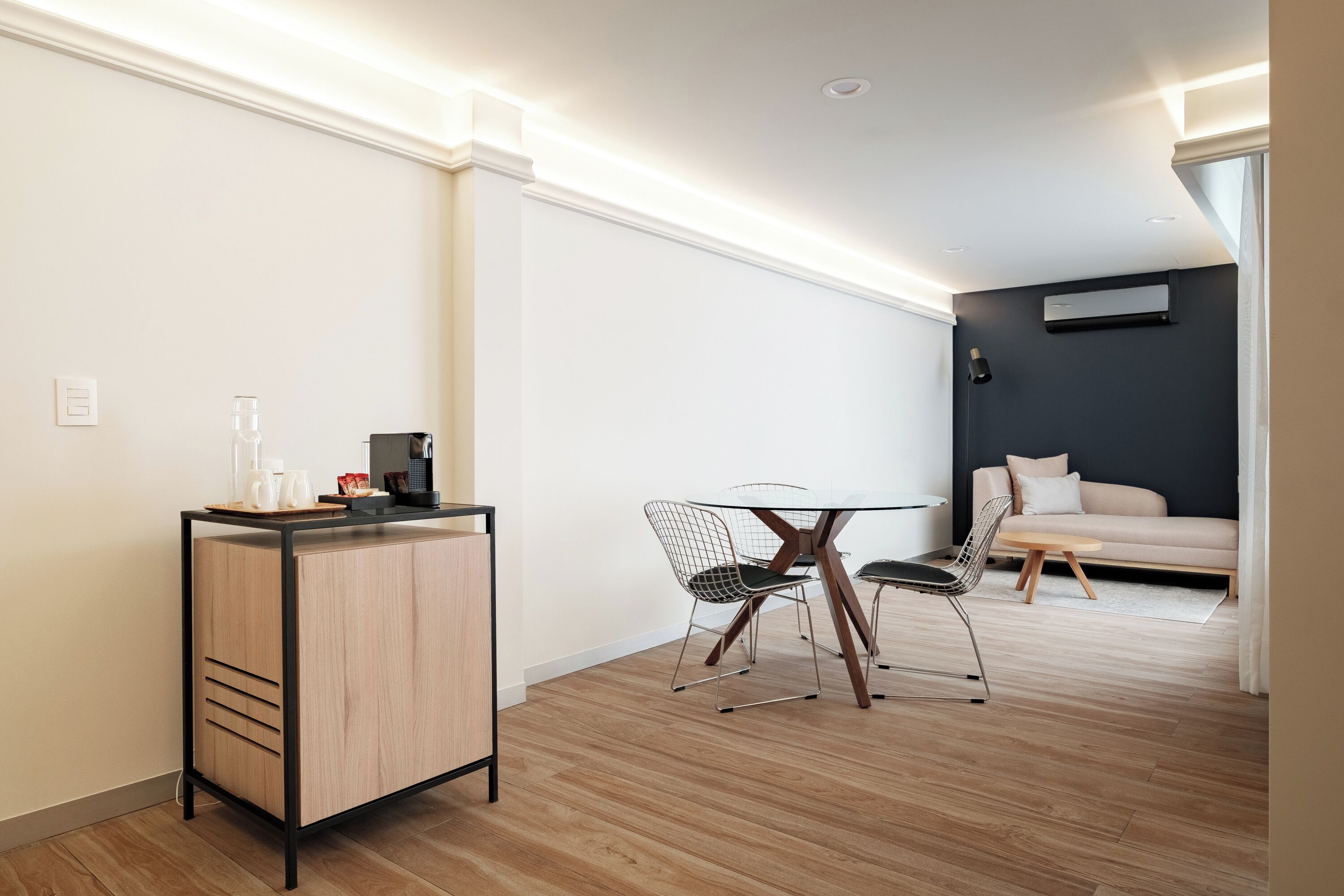 A modern living room with a dining area, featuring a wooden floor, a round table with wire chairs, a sofa, and a small cabinet with a coffee machine.