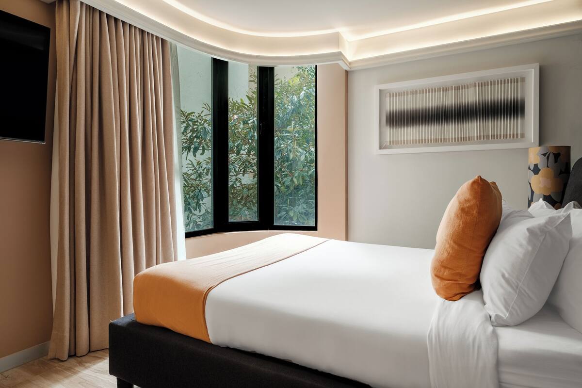 Een moderne hotelkamer met een groot bed, beige gordijnen, een raam met uitzicht op groen en een wandairconditioner.