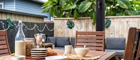 Un espacio exterior de comedor con una mesa de madera preparada para el desayuno, incluyendo tazones, tazas y una botella de leche.